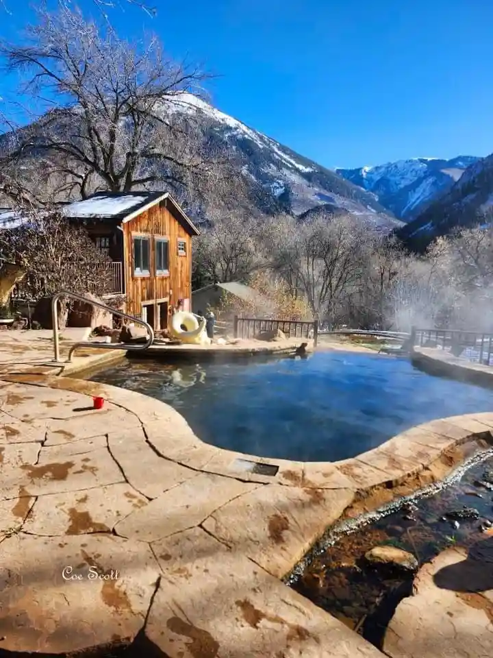 A scenic photo of a steaming outdoor hot tub surrounded by snow-dusted mountains and bare trees under a clear blue sky. A…