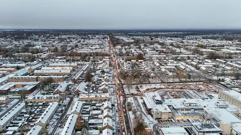An aerial view of a snow-covered suburban area with a long, dense traffic jam visible along a central road, stretching into…