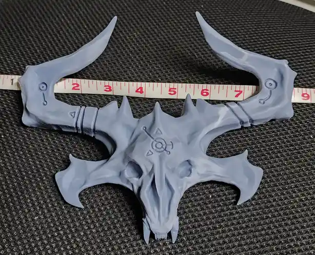 A light blue, skull-shaped, fantasy weapon with curved horns and spikes lies on a textured gray surface, with a measuring…