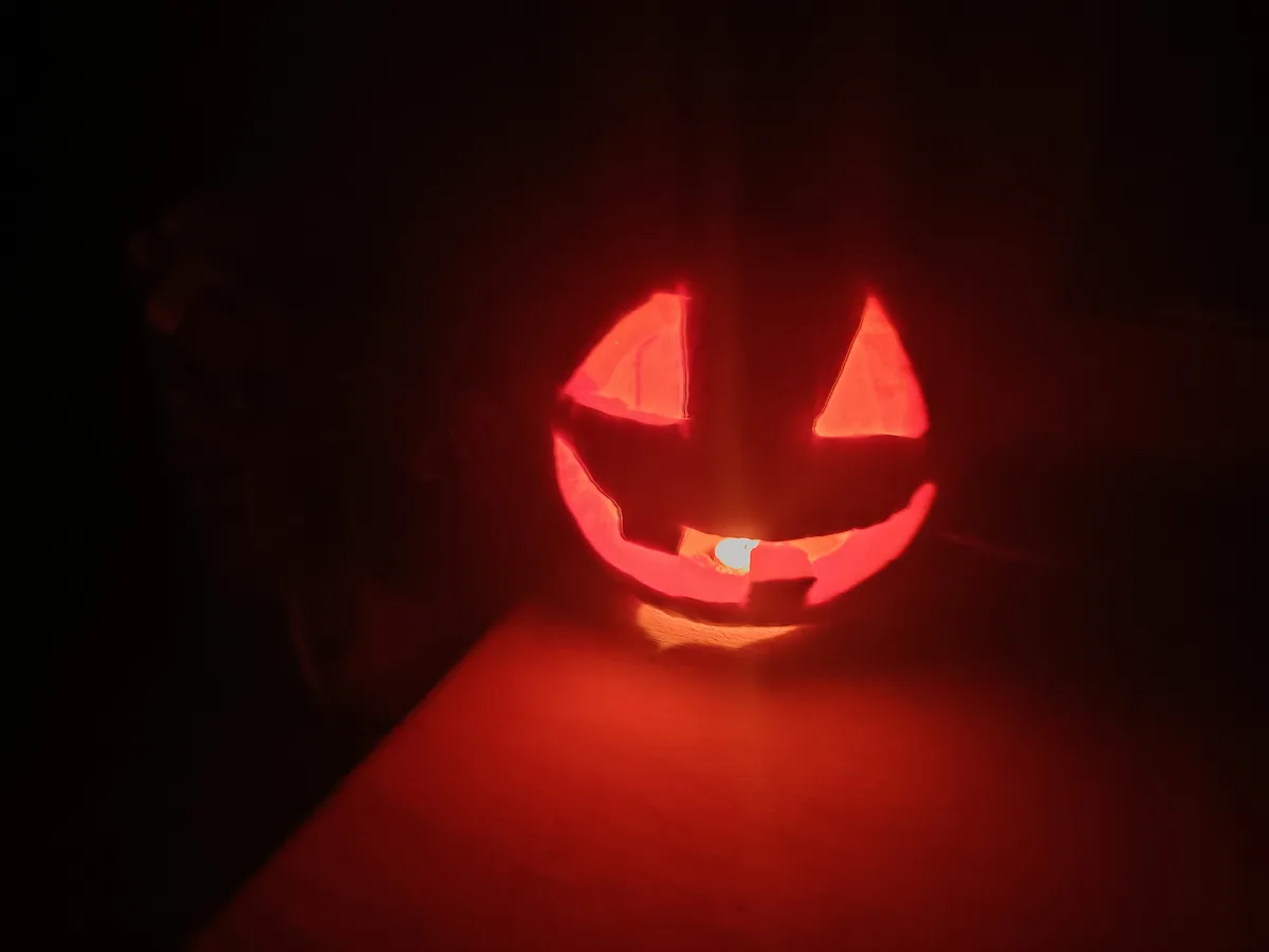A carved pumpkin with triangular eyes and a wide, toothy grin glows red in the dark, illuminated from within by a small…