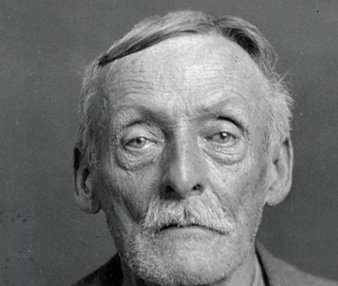 A black-and-white mugshot-style photo of an elderly man with a stern expression, thinning hair, and a mustache. Overlaid…