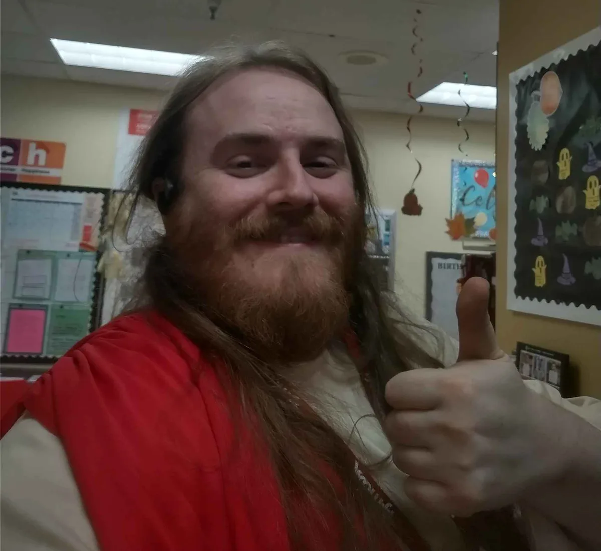 A man with long hair and a beard, wearing a red vest over a white shirt and a black earpiece, gives a thumbs-up in what…