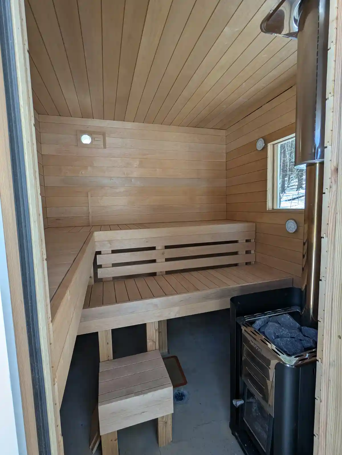An empty, newly built wooden sauna with benches, a Harvia heater, and a window showing snow outside. The overlaid text reads…
