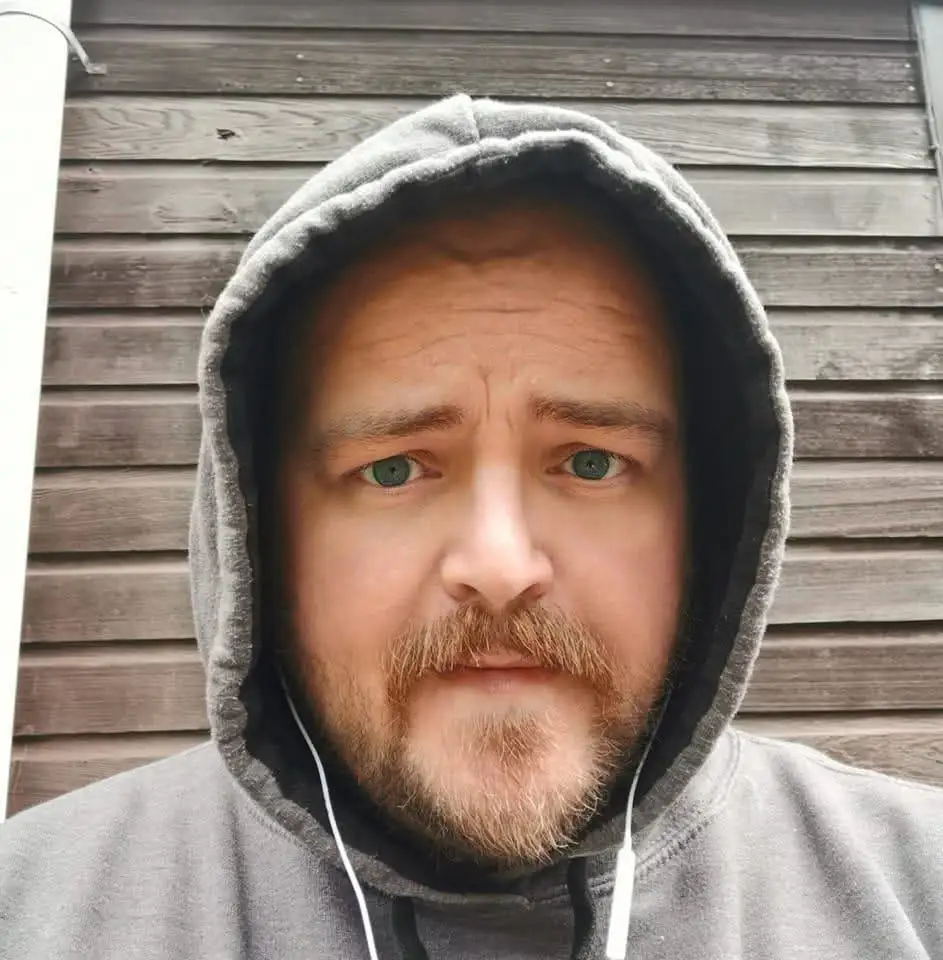 A man with a beard and green eyes wearing a grey hoodie with the hood up and white earbuds in his ears, standing in front of…