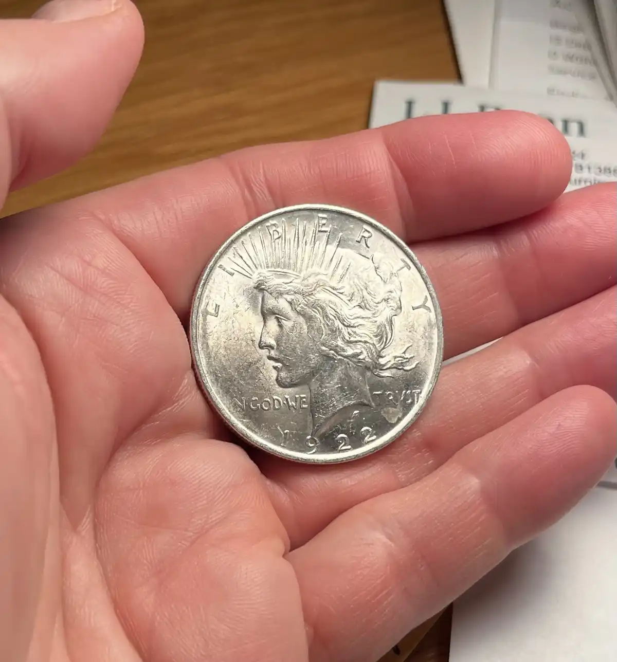 A close-up photo shows a person's hand holding a silver 1922 Peace Dollar coin, which clearly displays the profile of Lady…