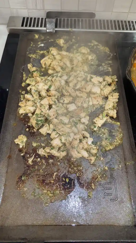 A close-up photo shows diced chicken pieces sizzling on a hot, greasy griddle with visible charred spots and green herb…