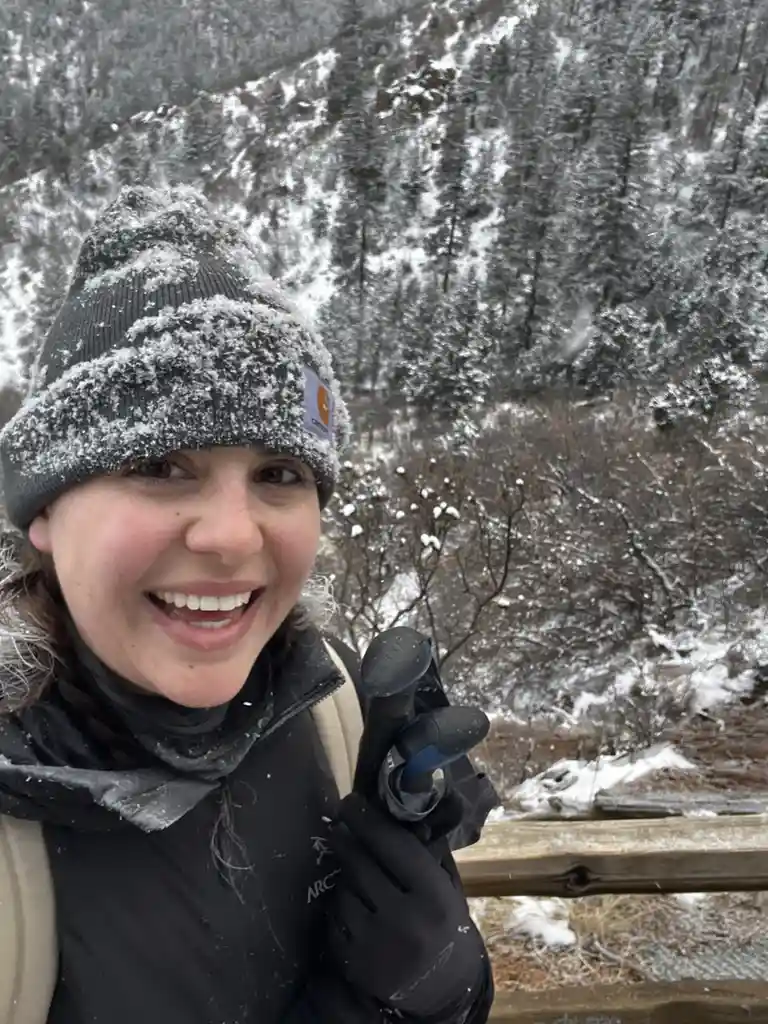 A person wearing a snow-dusted beanie and gloves smiles while holding trekking poles in a snowy forest, with text overlaid…