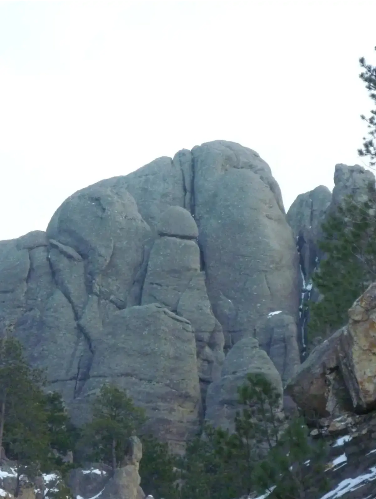 A photo of a large, gray rock formation with jagged edges and some snow patches, surrounded by evergreen trees under an…