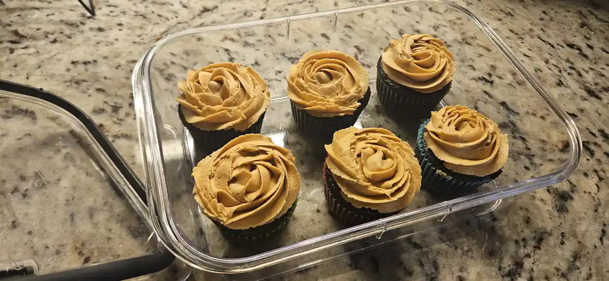 Five chocolate cupcakes with light brown frosting, arranged in a clear plastic container on a speckled countertop. The…