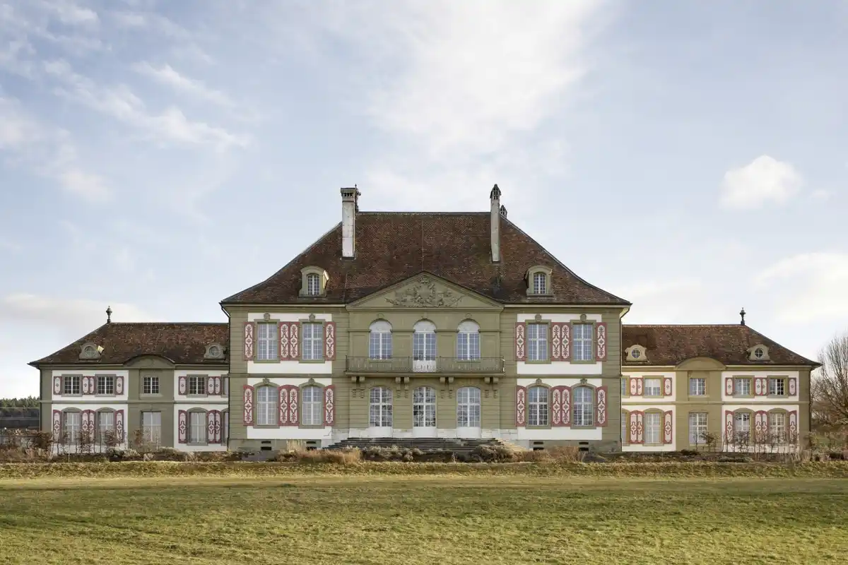 a large, ornate, multi-story house with a brown roof and decorative red-and-white shutters stands in a grassy field under a…