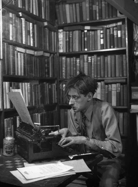 A black-and-white photo of a man with glasses and messy hair, sitting at a vintage typewriter in a room surrounded by…