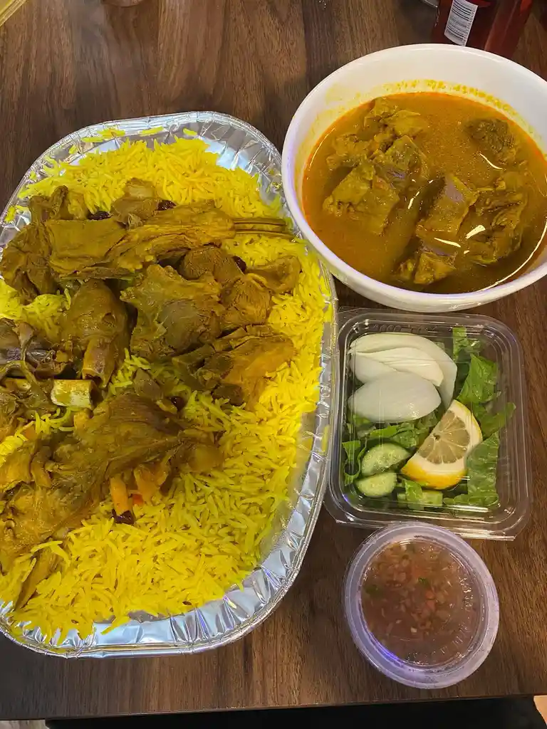 A meal featuring a foil tray of yellow rice topped with meat, a bowl of curry, a container of salad with onions, cucumber…