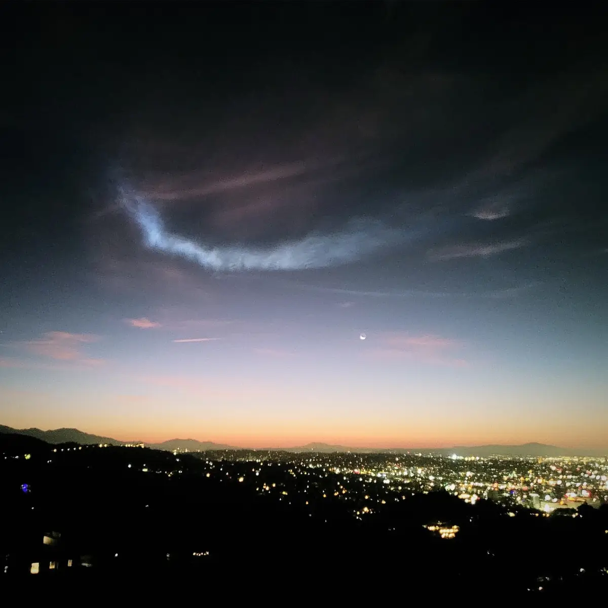 A nighttime cityscape photo shows a crescent moon in a twilight sky above a sprawling urban area with lights. A large, wispy…