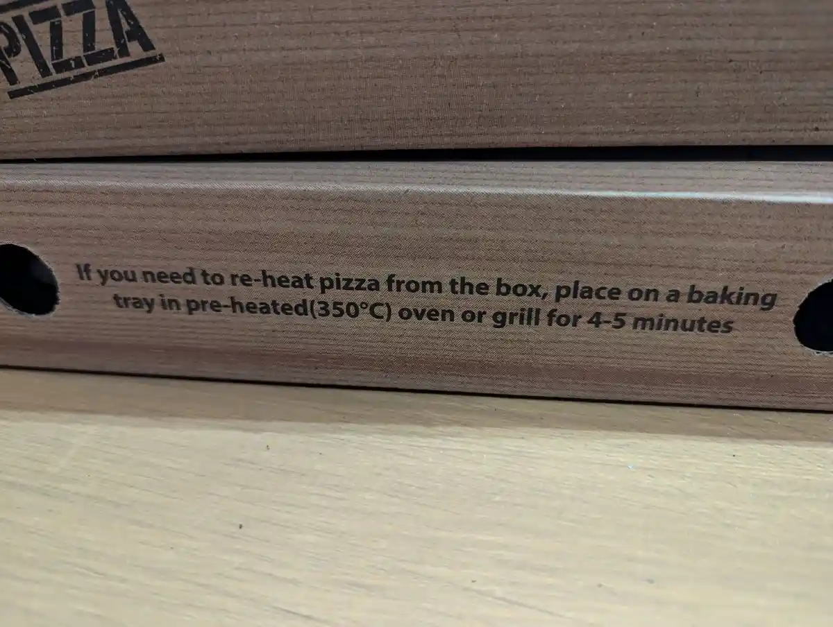 A close-up of a brown cardboard pizza box with printed text instructing users to reheat pizza from the box by placing it on…