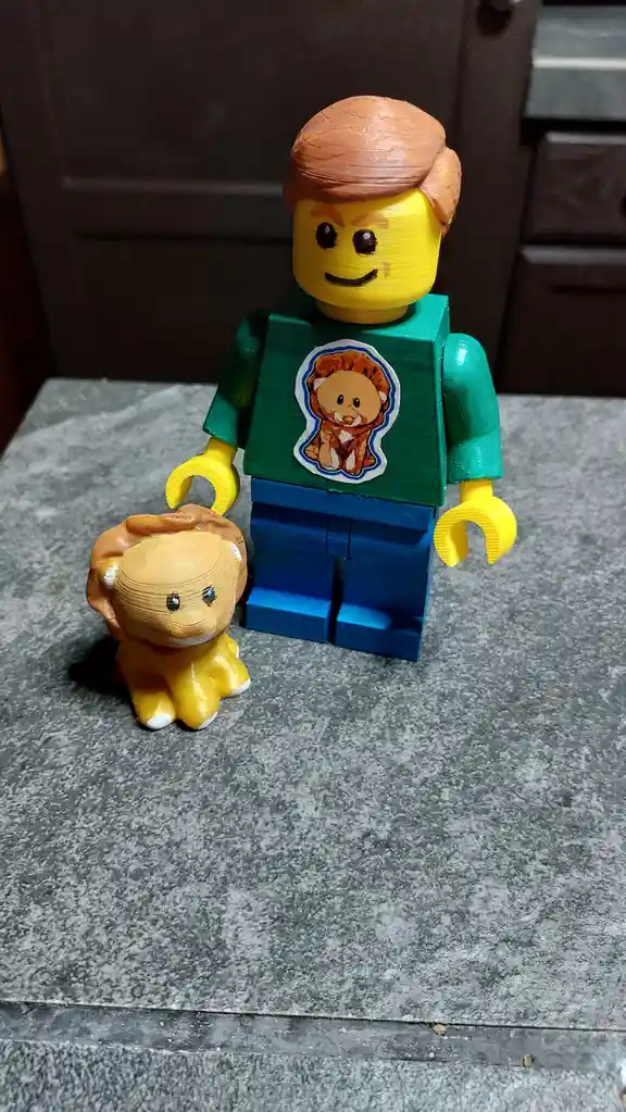 A 3d-printed lego minifigure with brown hair, wearing a green shirt with a lion sticker and blue pants, stands next to a…
