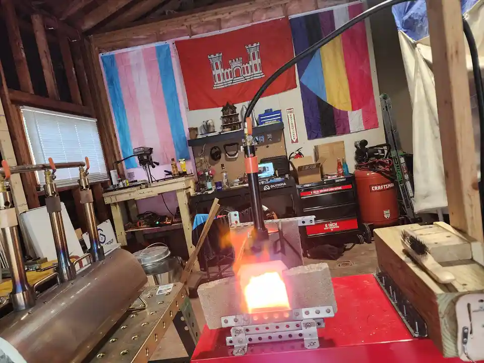 A workshop with a torch heating a metal block, surrounded by tools and three flags: a trans flag, a Colorado state flag, and…