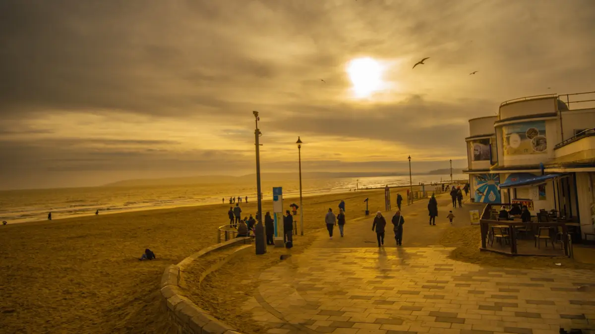 A golden-hued beach scene at sunset with people walking along a promenade and sitting on the sand, overlaid with the text…