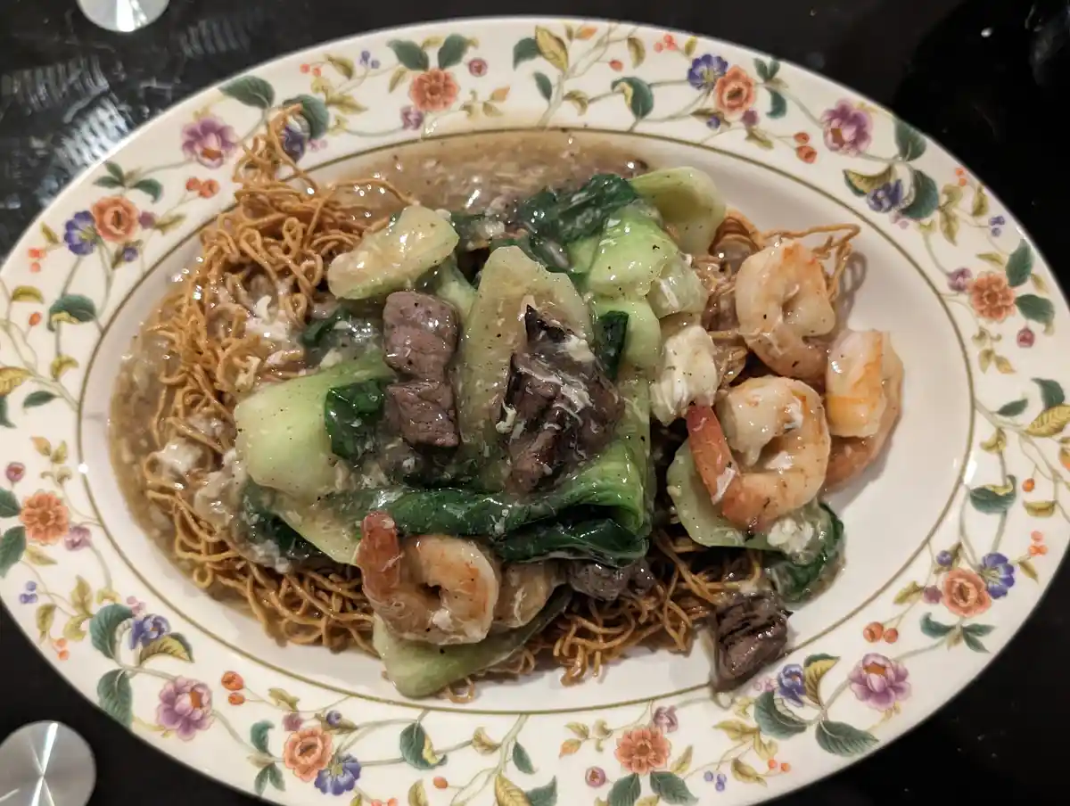 A close-up photo shows a plate of stir-fried noodles topped with shrimp, beef, and bok choy, served on a decorative…