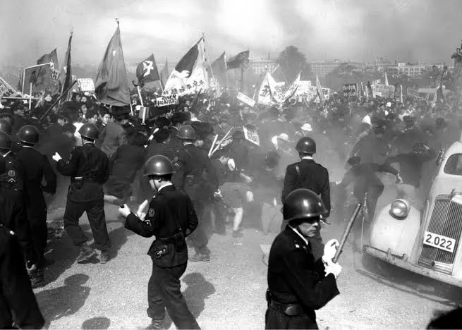 A black-and-white historical photograph shows a large crowd of protesters holding flags and signs, facing off against…