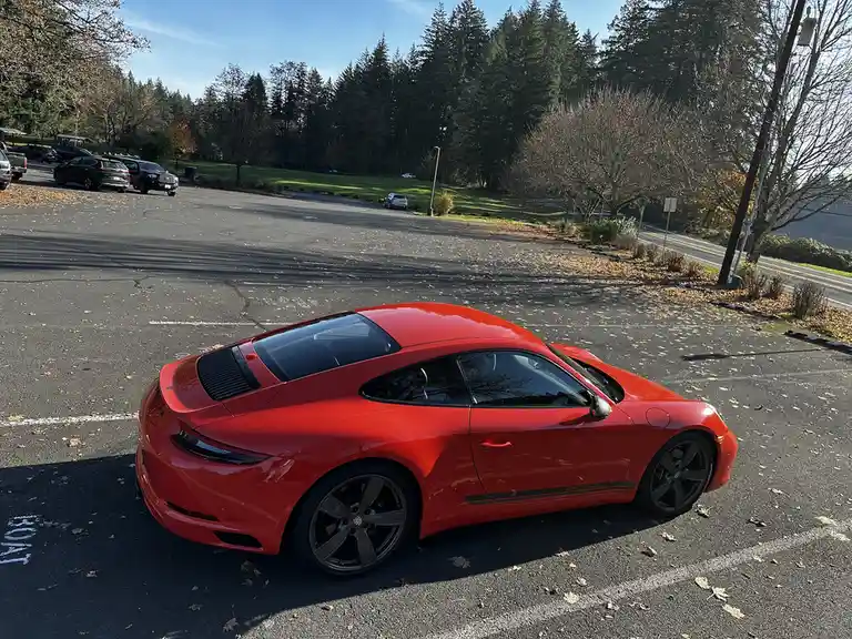 A red Porsche 911 Carrera is parked in an asphalt lot with white painted lines, occupying a space marked with the word…
