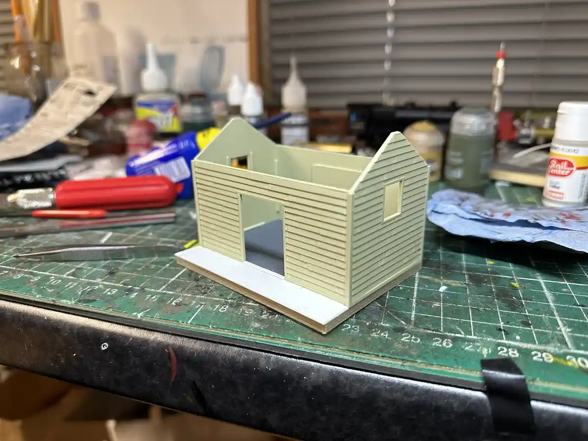 A small, unpainted model house with a gabled roof and open doorway sits on a green gridded work mat surrounded by hobby…