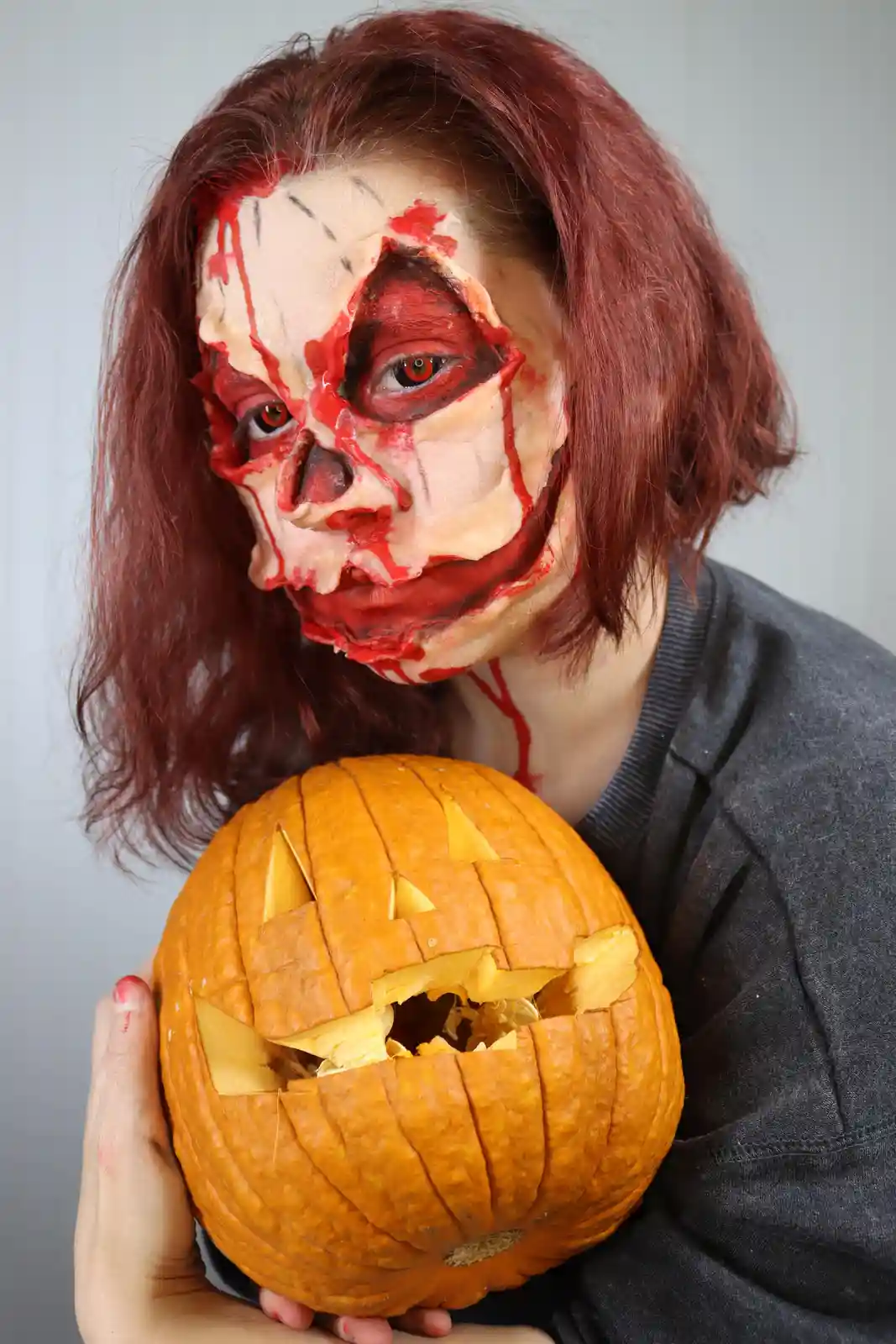 A person with elaborate zombie makeup, featuring fake blood and torn skin effects, holds a carved jack-o'-lantern. The…