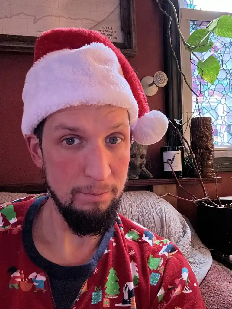 A man with a beard wears a red and white Santa hat and festive red pajamas with Christmas-themed patterns. He stares…