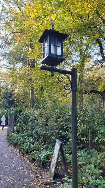 A black ornate lamppost stands on a brick path in a leafy park, with a small A-frame sign at its base reading 'Snoepwinkel…