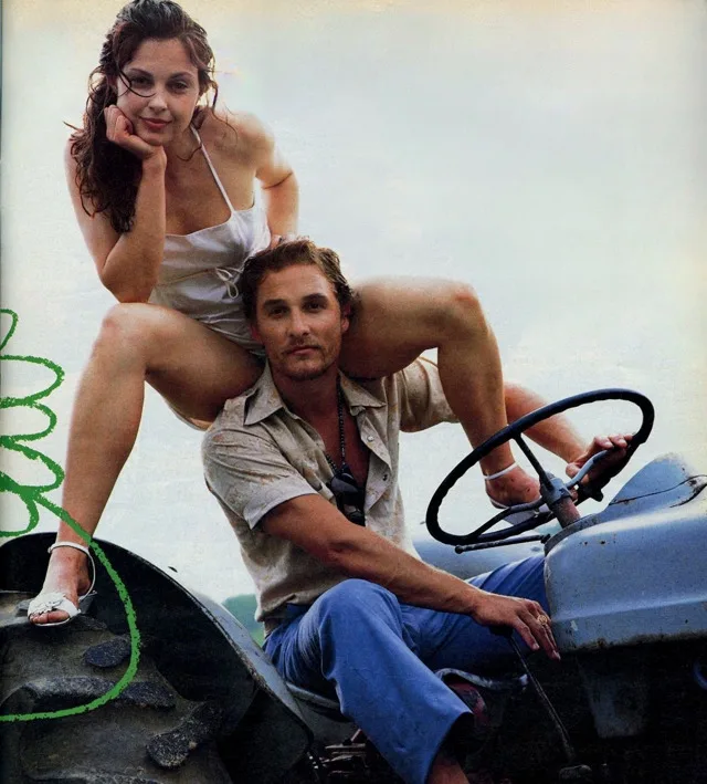 A man sits in the driver's seat of a blue tractor, holding the wheel, while a woman in a white top sits on his shoulders. A…