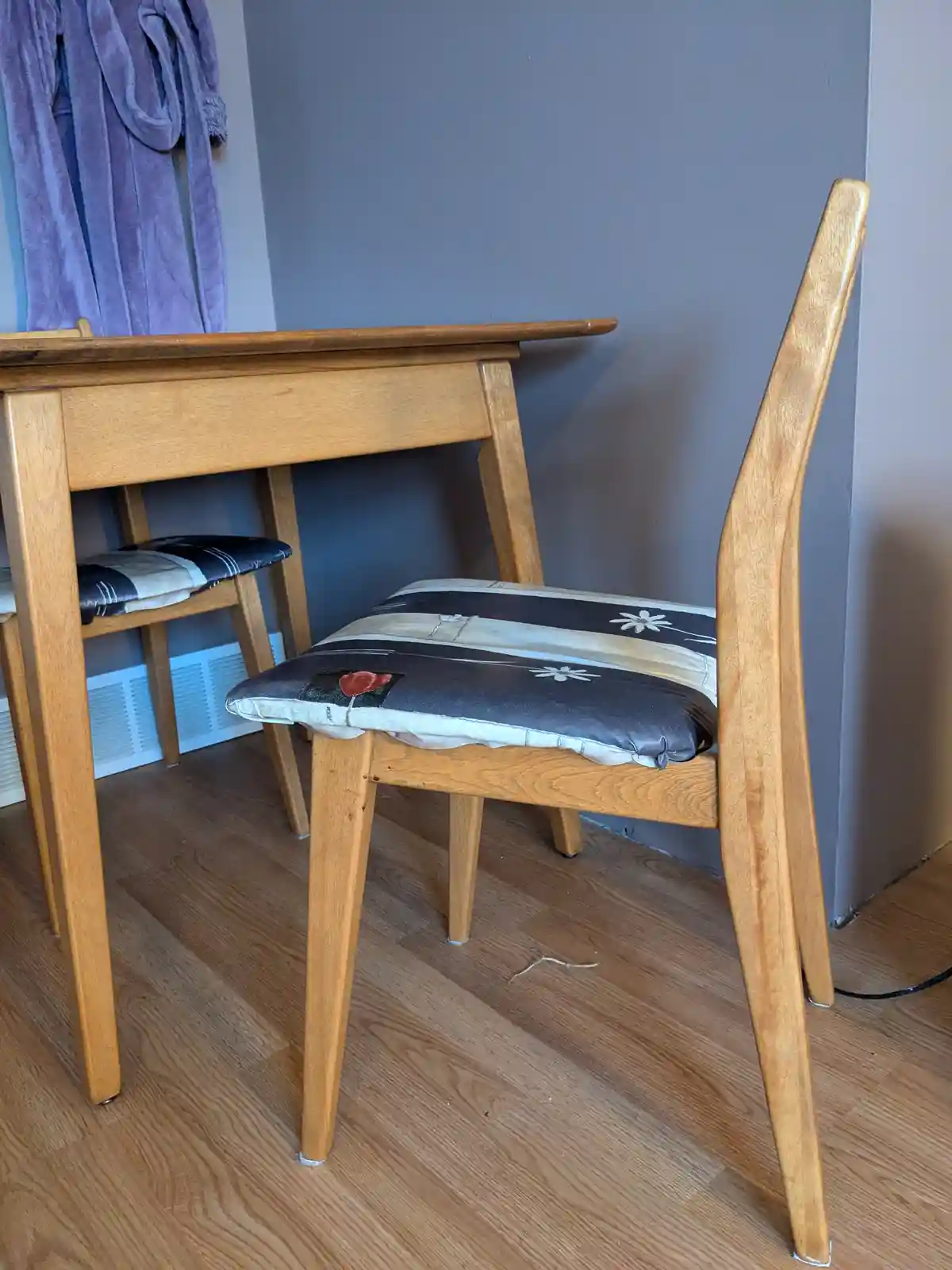 A wooden dining chair with a patterned cushion sits at a matching wooden table on a hardwood floor, with a purple robe…