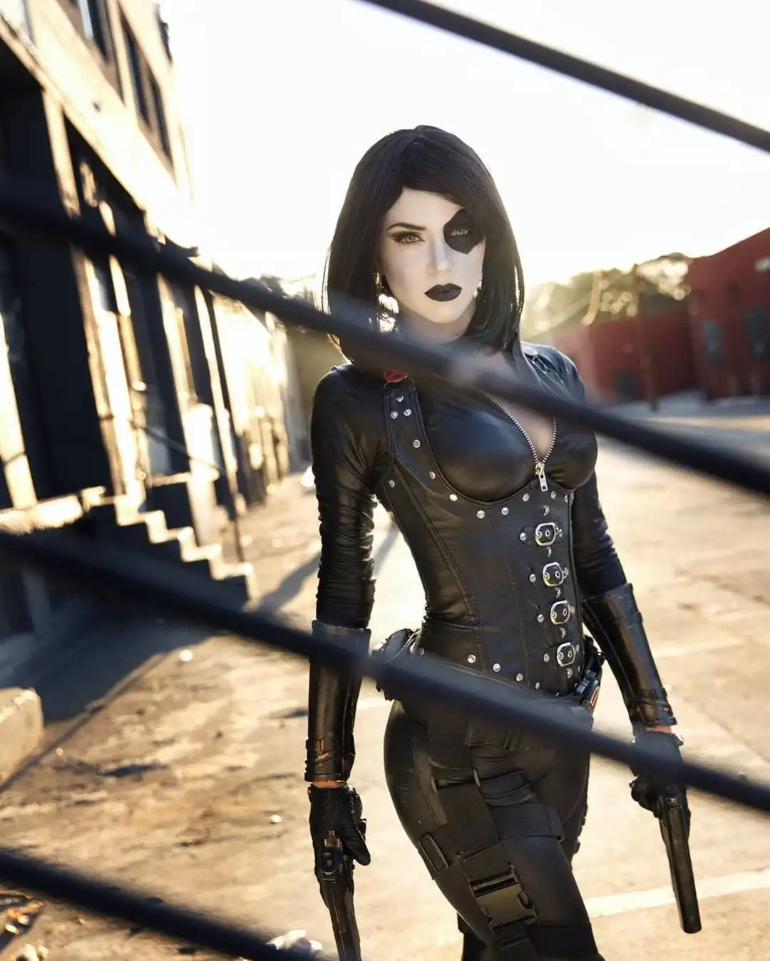 A person in a black leather corset and pants, with a black eye patch over one eye and dark lipstick, stands in an urban…