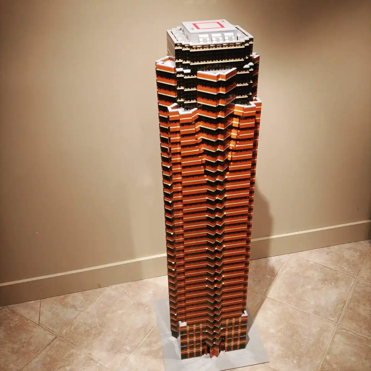 A tall, intricate Lego model of a skyscraper with orange and black bricks stands on a tiled floor against a beige wall. The…