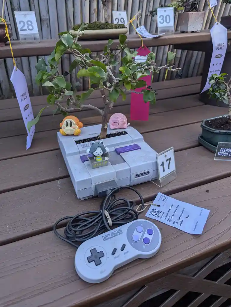 A Super Nintendo Entertainment System console is repurposed as a planter for a bonsai tree, with small crochet plushies…