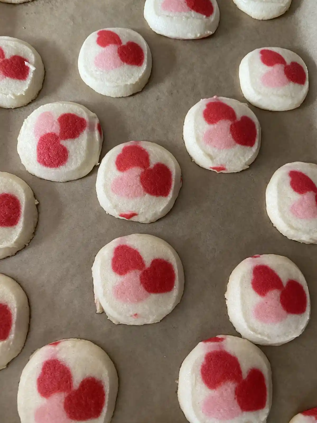 An overhead shot shows numerous round, white sugar cookies arranged on brown parchment paper. Each cookie is decorated with…