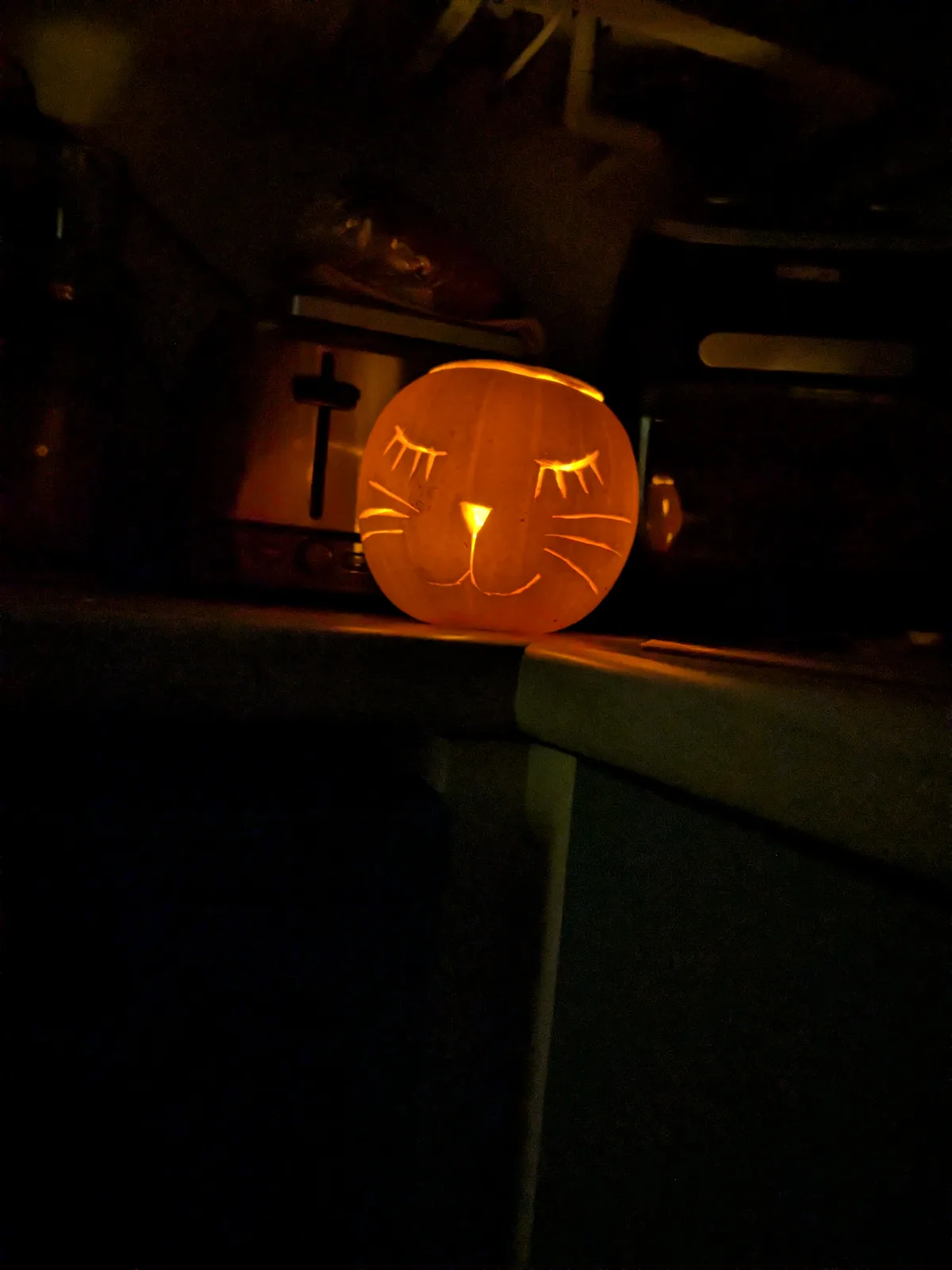 A carved pumpkin with cat-like features—long eyelashes, whiskers, and a triangular nose—is lit from within and sits on a…