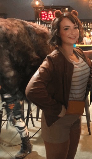 A woman with small brown bear ears on her head smiles at the camera while standing next to a large, furry, costumed bear…