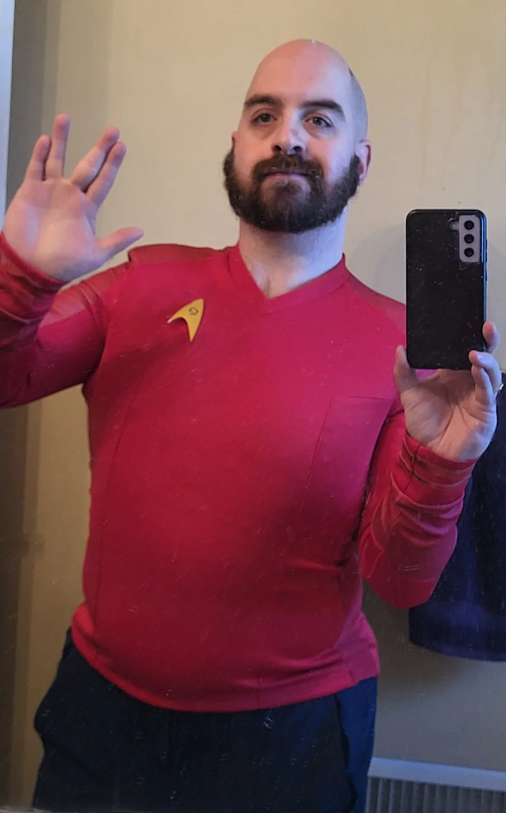 A bald man with a beard takes a mirror selfie while wearing a red Star Trek uniform with a gold insignia and holding a black…