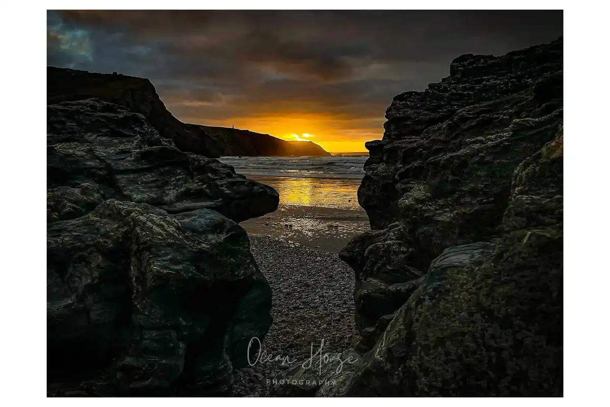 A dramatic photograph shows a sunset viewed through a narrow cleft between large, dark, textured rocks. The sun is low on…