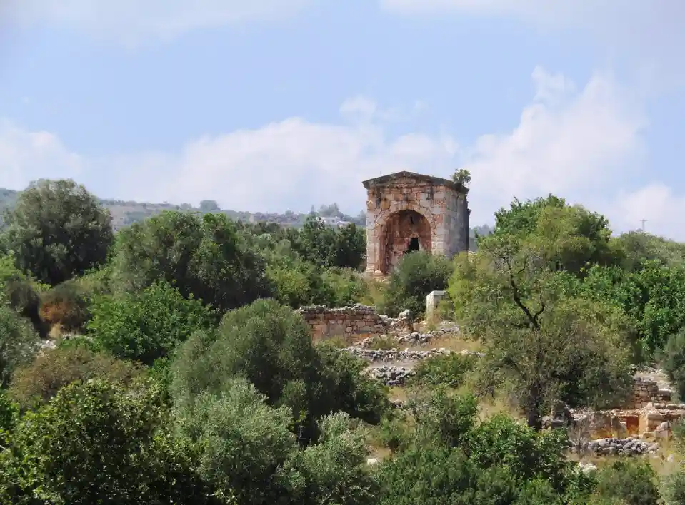 An ancient stone ruin with an arched doorway stands partially obscured by green trees and shrubs on a hillside under a…