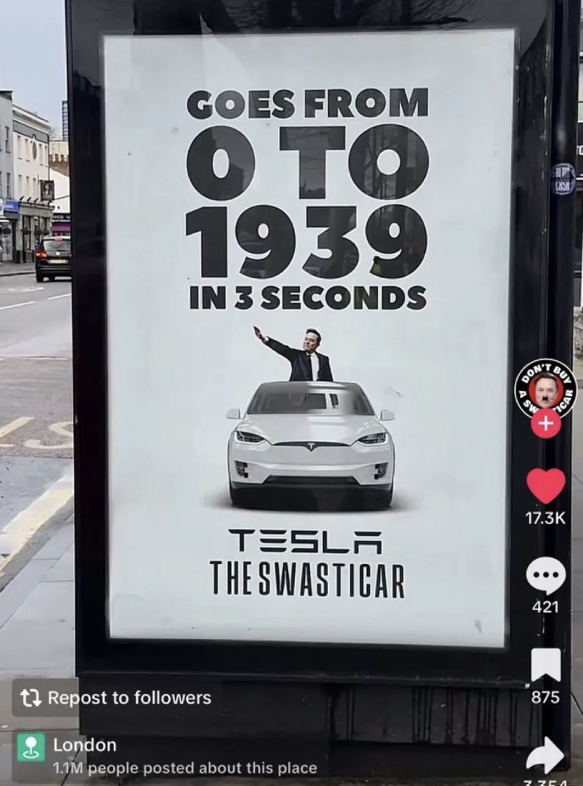 A street advertisement for a Tesla car features a man in a suit waving from the roof of the vehicle. The ad text claims the…