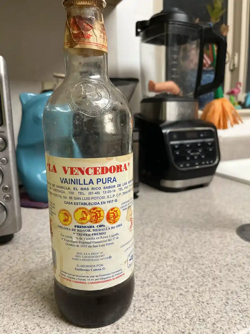 A close-up photo of an old, dusty glass bottle of 'La Vencedora Vainilla Pura' vanilla extract sitting on a kitchen counter…