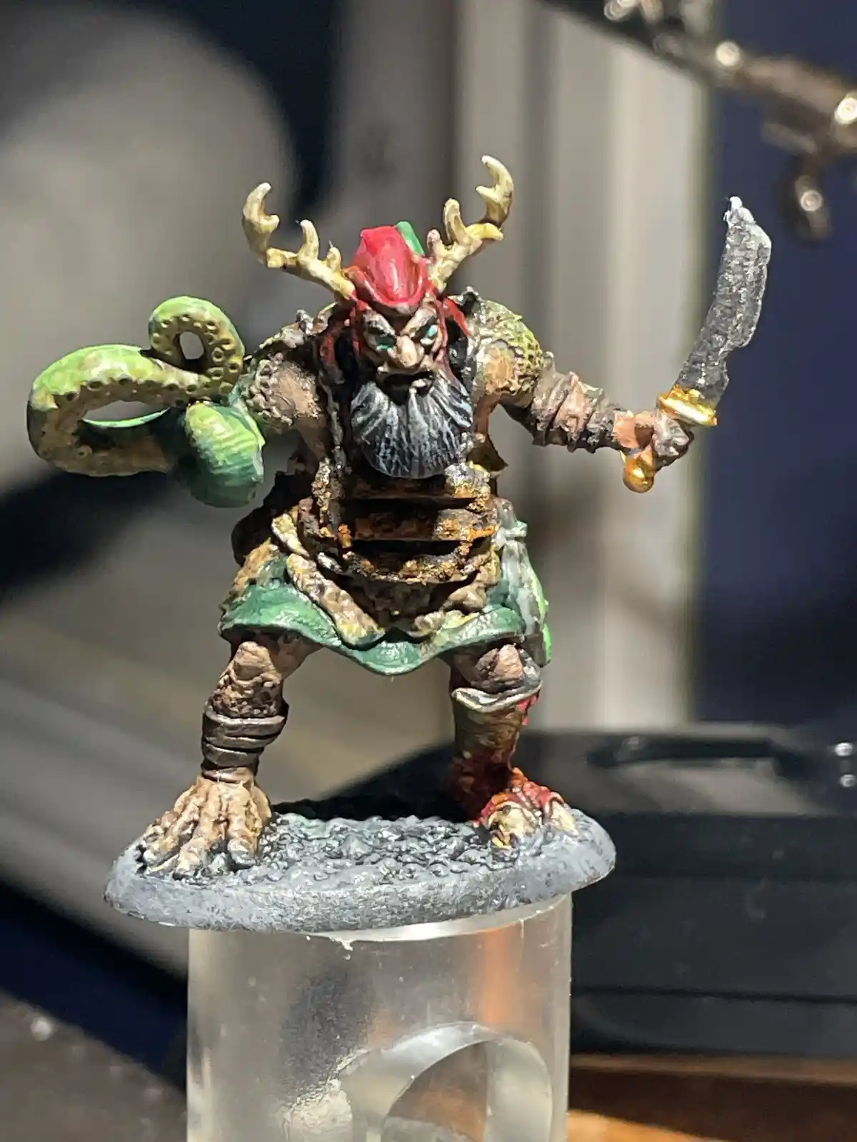 A hand-painted fantasy miniature of a stout, bearded creature with antlers, a red hat, and a green octopus tentacle on its…