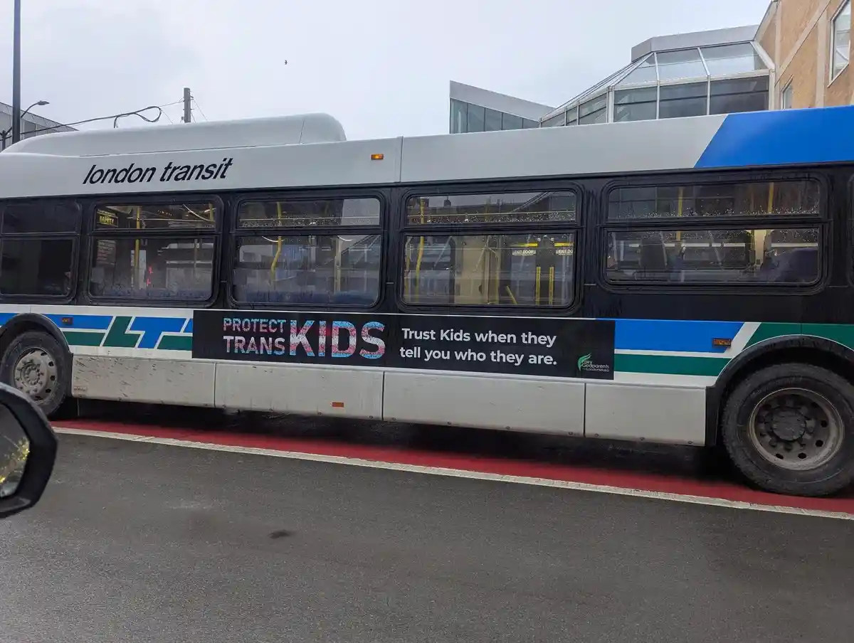 A London Transit bus displays a public service announcement banner that reads 'PROTECT TRANS KIDS' in large, colorful…
