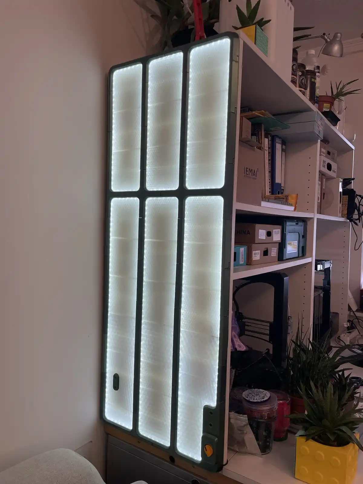 A tall, modular LED light panel with six glowing rectangular sections is mounted vertically against a wall next to a white…