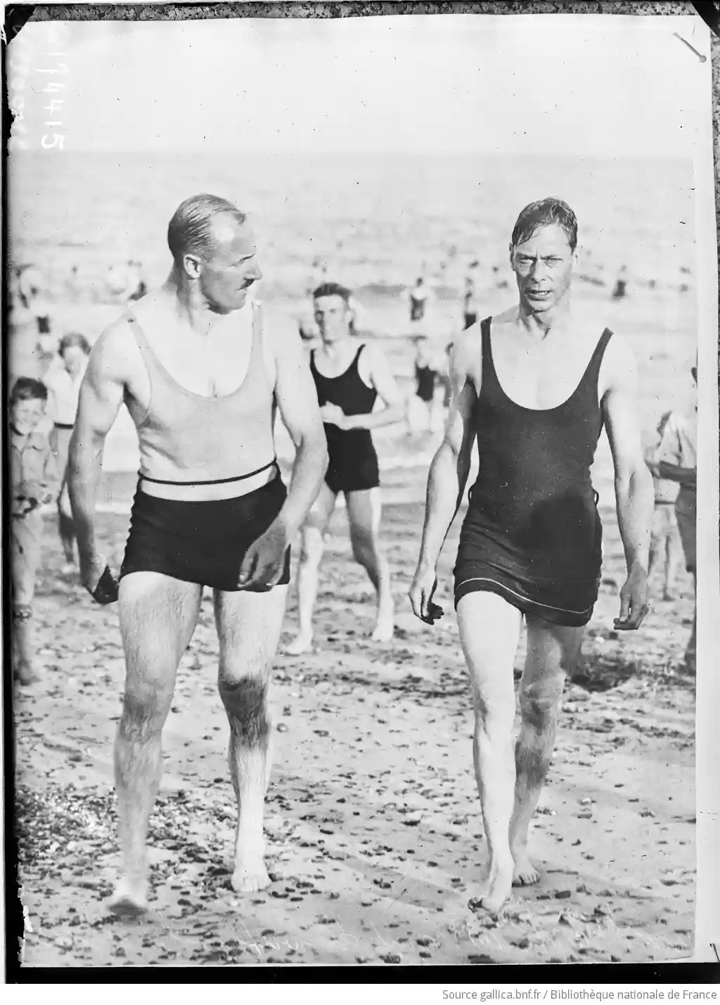 A vintage black-and-white photo shows two men in early 20th-century swimwear walking on a pebbly beach. The man on the left…