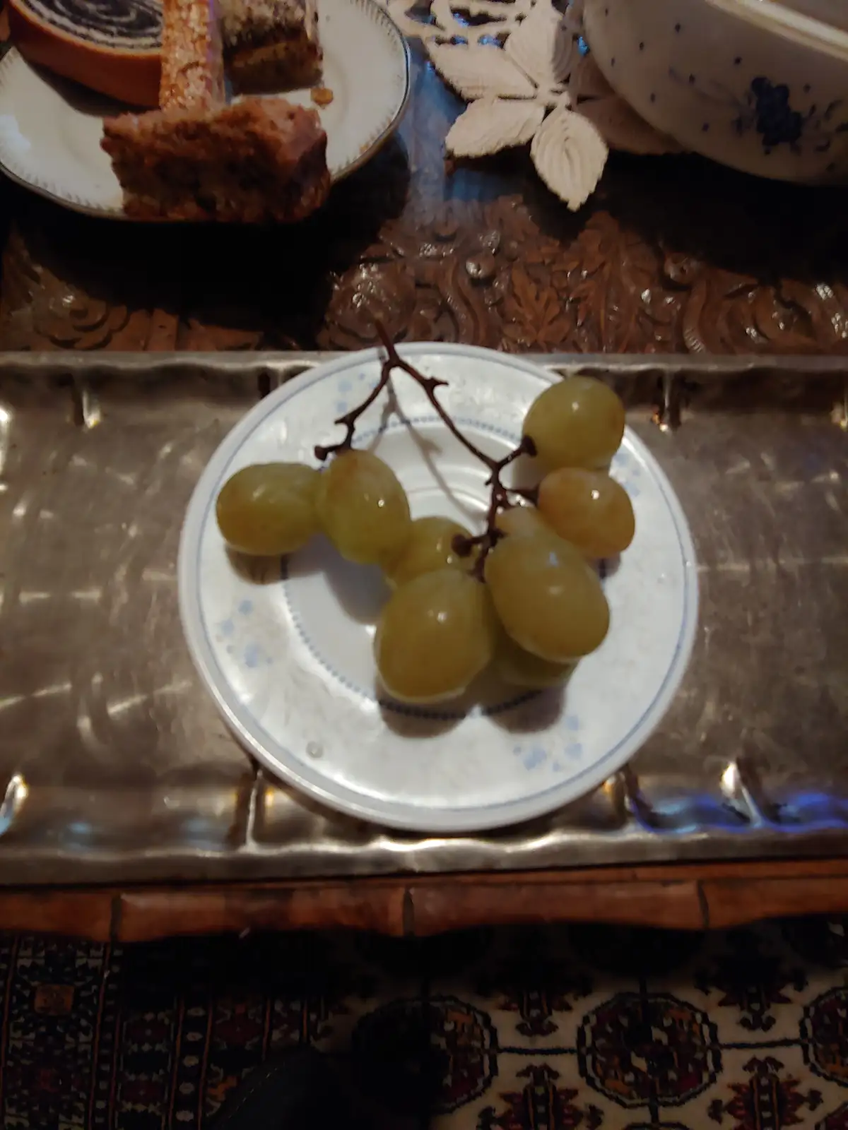 A close-up photo shows a small bunch of green grapes on a white plate with blue floral patterns, sitting on a silver tray…