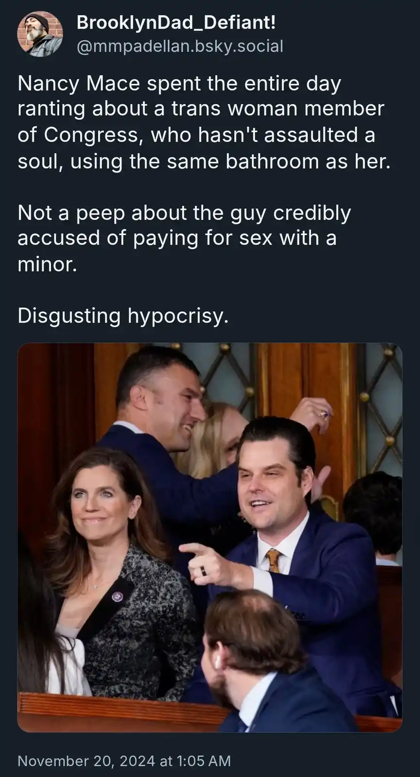 The image shows a woman in a patterned blazer and a man in a blue suit pointing and smiling in what appears to be a…