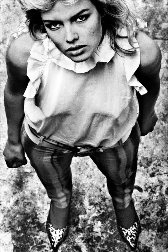 A high-angle black-and-white photo shows a woman looking down intensely at the camera, wearing a ruffled top, jeans, and…