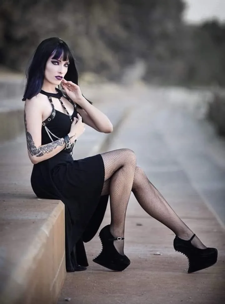 A woman with dark hair and gothic makeup sits on a concrete ledge outdoors, wearing a black dress with straps, fishnet…