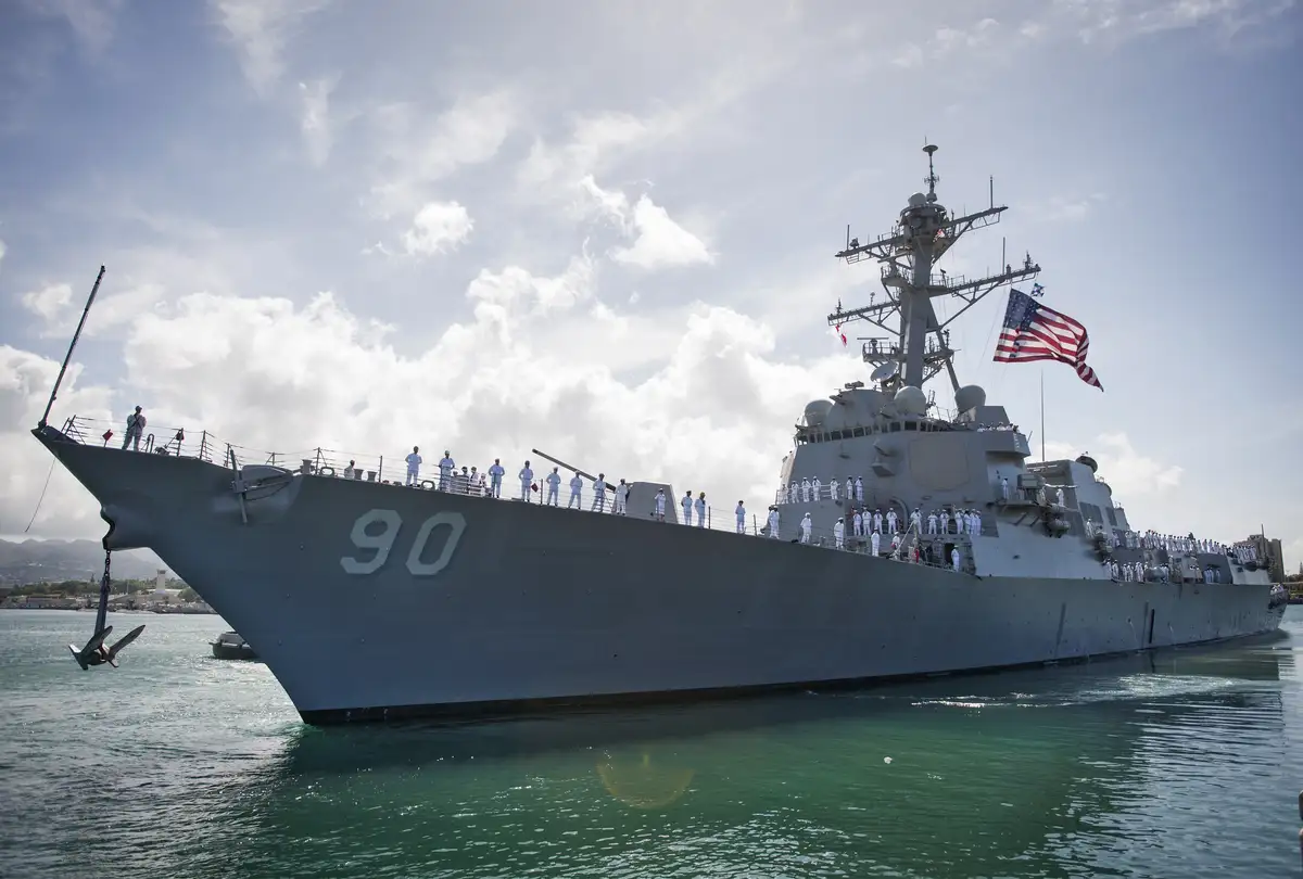 A large gray U.S. Navy destroyer, hull number 90, is docked in calm green water under a partly cloudy sky. Numerous sailors…